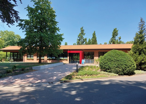 1-geschossiges Gebäude der Institutsambulanz mit hellbraunem Dach und mit rot umrahmter Eingangstür, eingebettet in grünen Klinikpark.