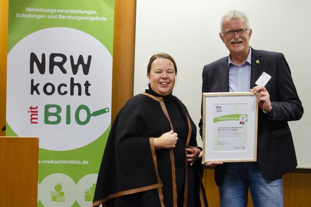 Eine Frau und ein Mann stehen vor dem Plakat "NRW koch Bio" und halten ein Zertifikat.